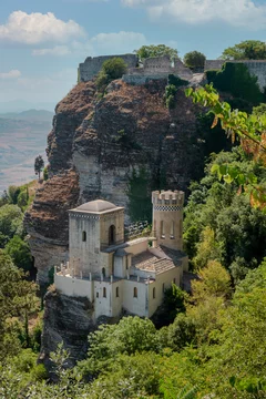 Castillo medieval en la cima de un acantilado rodeado de vegetación, con el paisaje montañoso de Sicilia al fondo